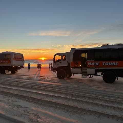 Tour buses parked on a beach at sunset with people nearby.