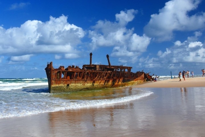 Rusty shipwreck on a sandy beach with people nearby under a blue sky.