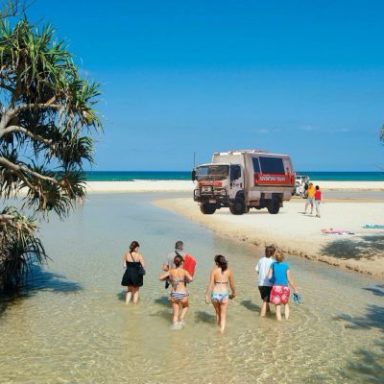 People walking in shallow water towards an off-road vehicle on a sandy beach.