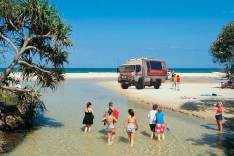 People walking in shallow water towards an off-road vehicle on a sandy beach.