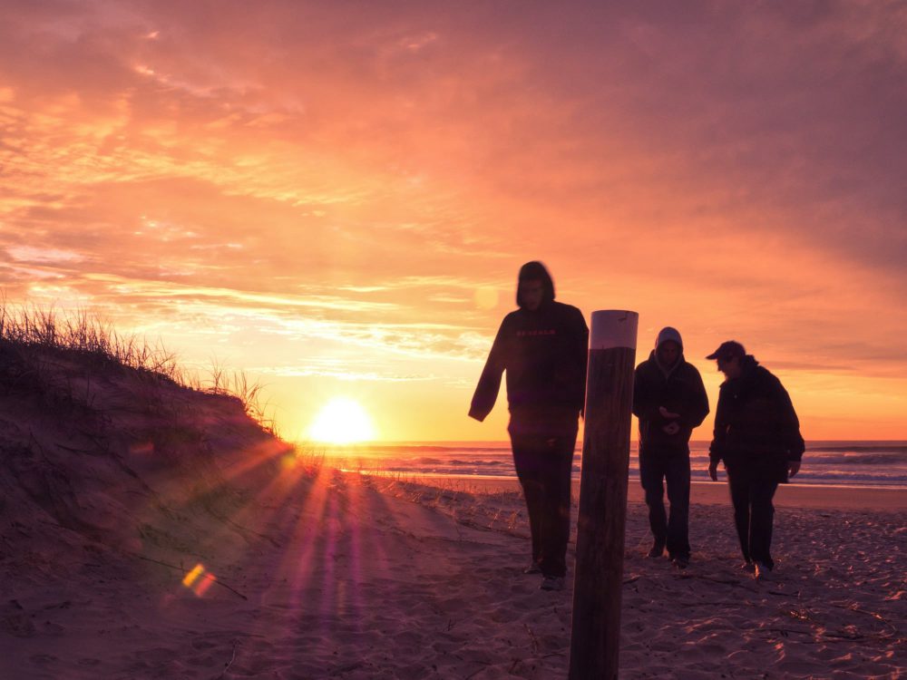 Three people walking on a beach at sunrise with colorful sky.