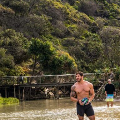 Man standing in shallow water near a boardwalk and greenery.