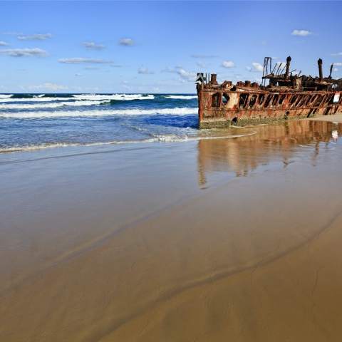 Rusty shipwreck on a sandy beach with waves and a clear sky.