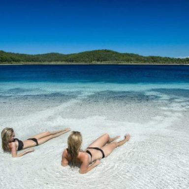 Two people sunbathing on a white sandy beach by a clear blue lake.