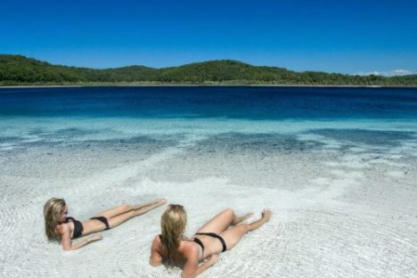 Two people sunbathing on a white sandy beach by a clear blue lake.