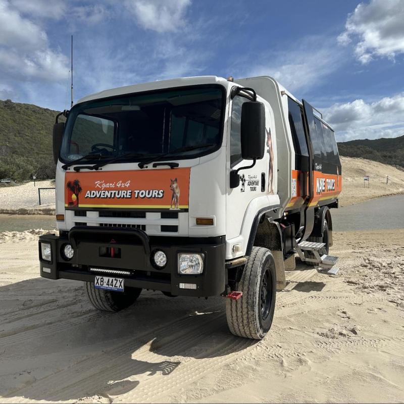 Tour truck on sandy terrain with hills and water in background.